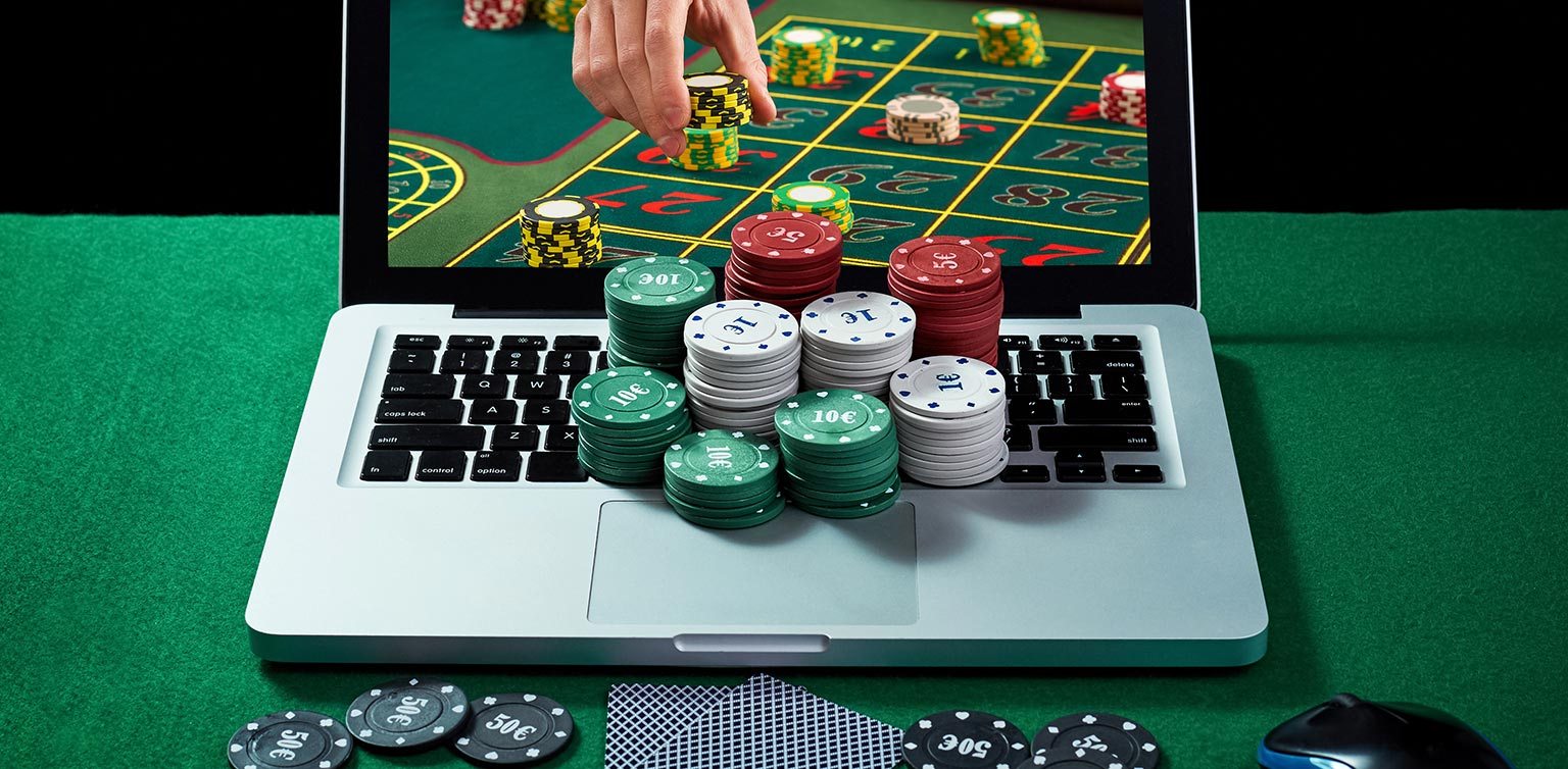 A photo of a laptop, with chips stacks on the keyboard, displaying a man’s hand placing chips on a roulette layout