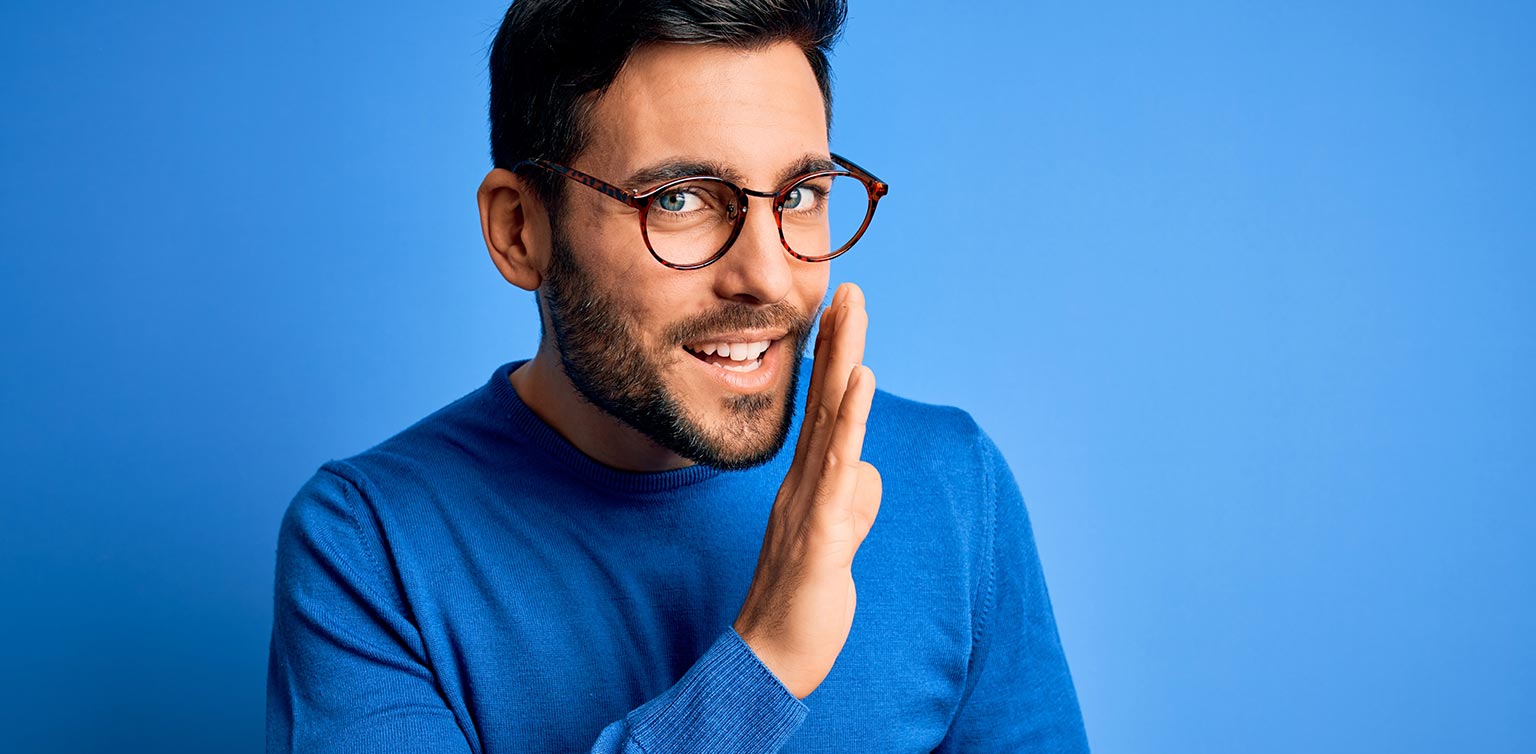 A young bearded guy wearing glasses and a blue sweater sharing a secret with the view