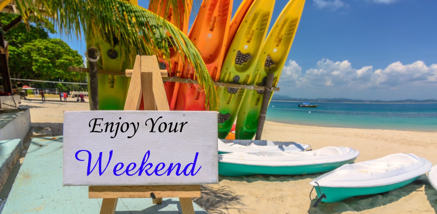 A photo of a tropical beach with kayaks and a white canvas sign reading ‘enjoy your weekend’