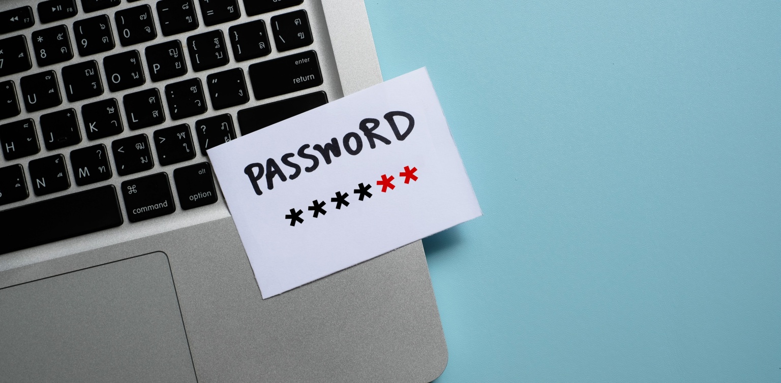 An image of a grey laptop with a white posted note displaying a password in stars on a blue surface 