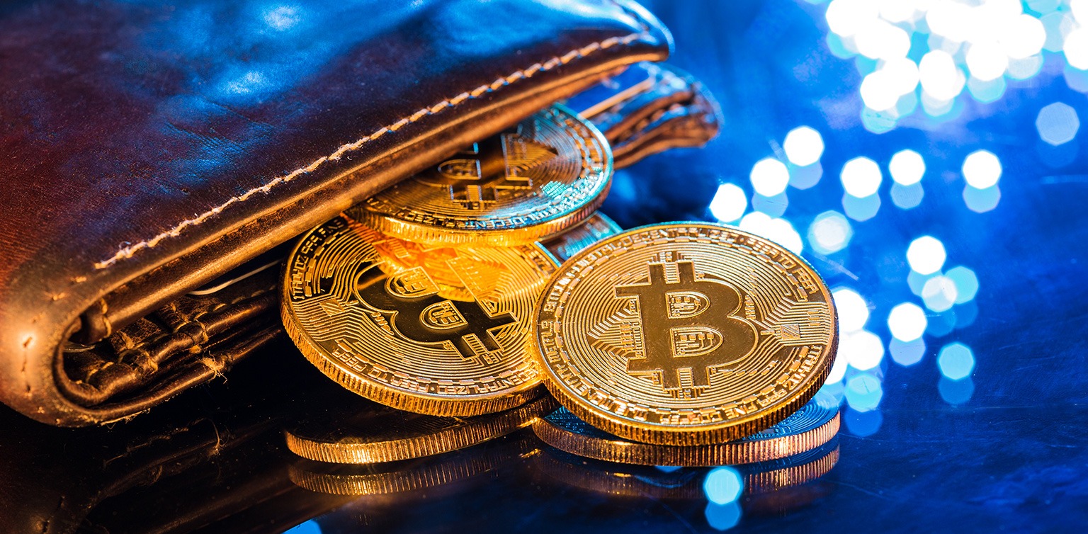 A close-up photograph of gold Bitcoin tokens spilling out of a brown leather wallet on a blue background