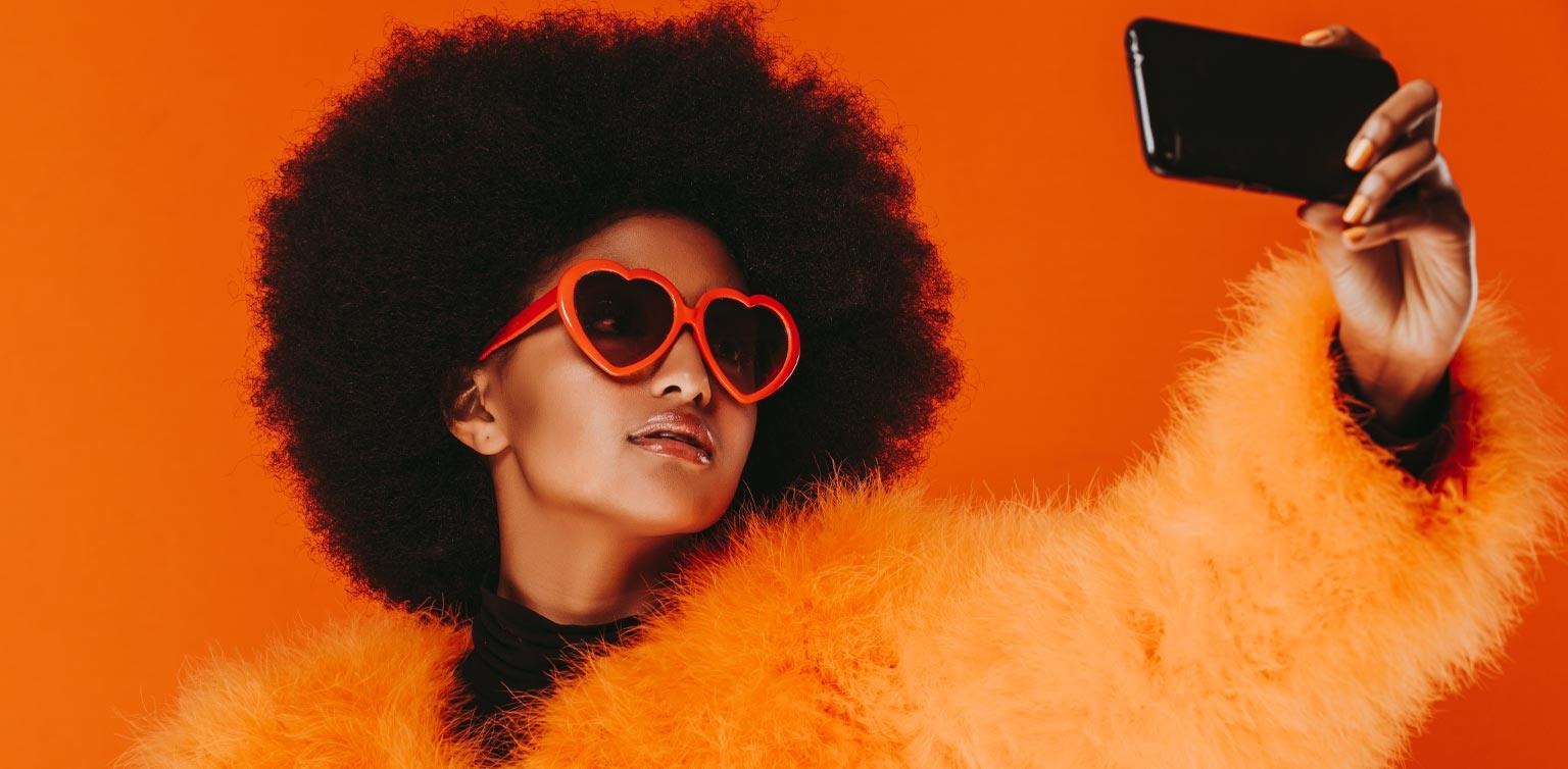 A woman wearing faux fur and heart-shaped dark glasses taking selfies on her smartphone