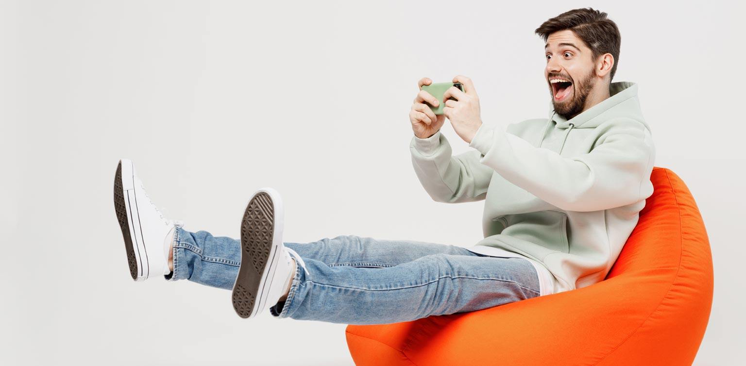 A delighted man sitting on an orange pouf with his legs outstretched playing games on his phone