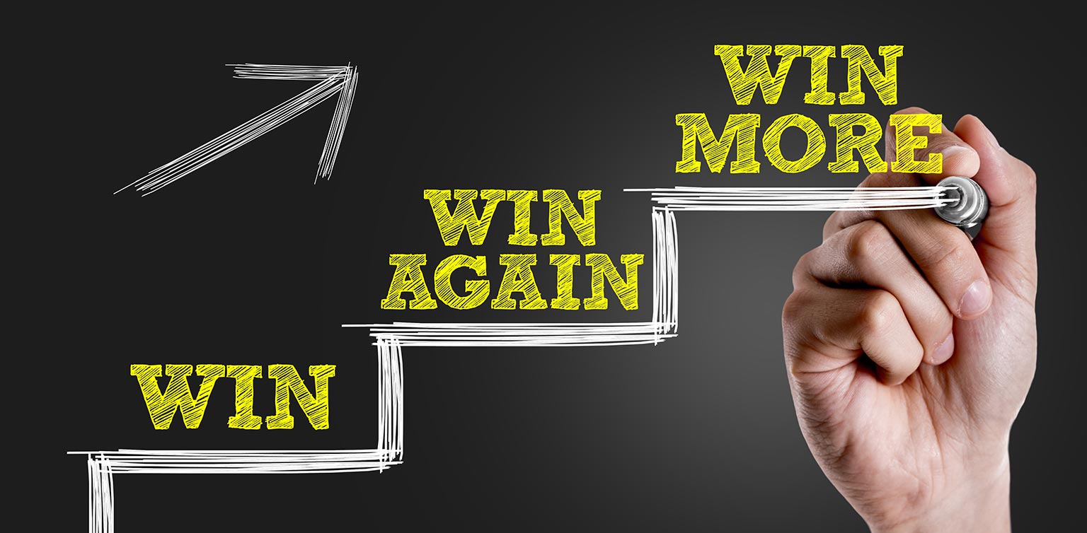 A photo of a hand writing Win, Win Again and Win More on a three-step configuration on a black background