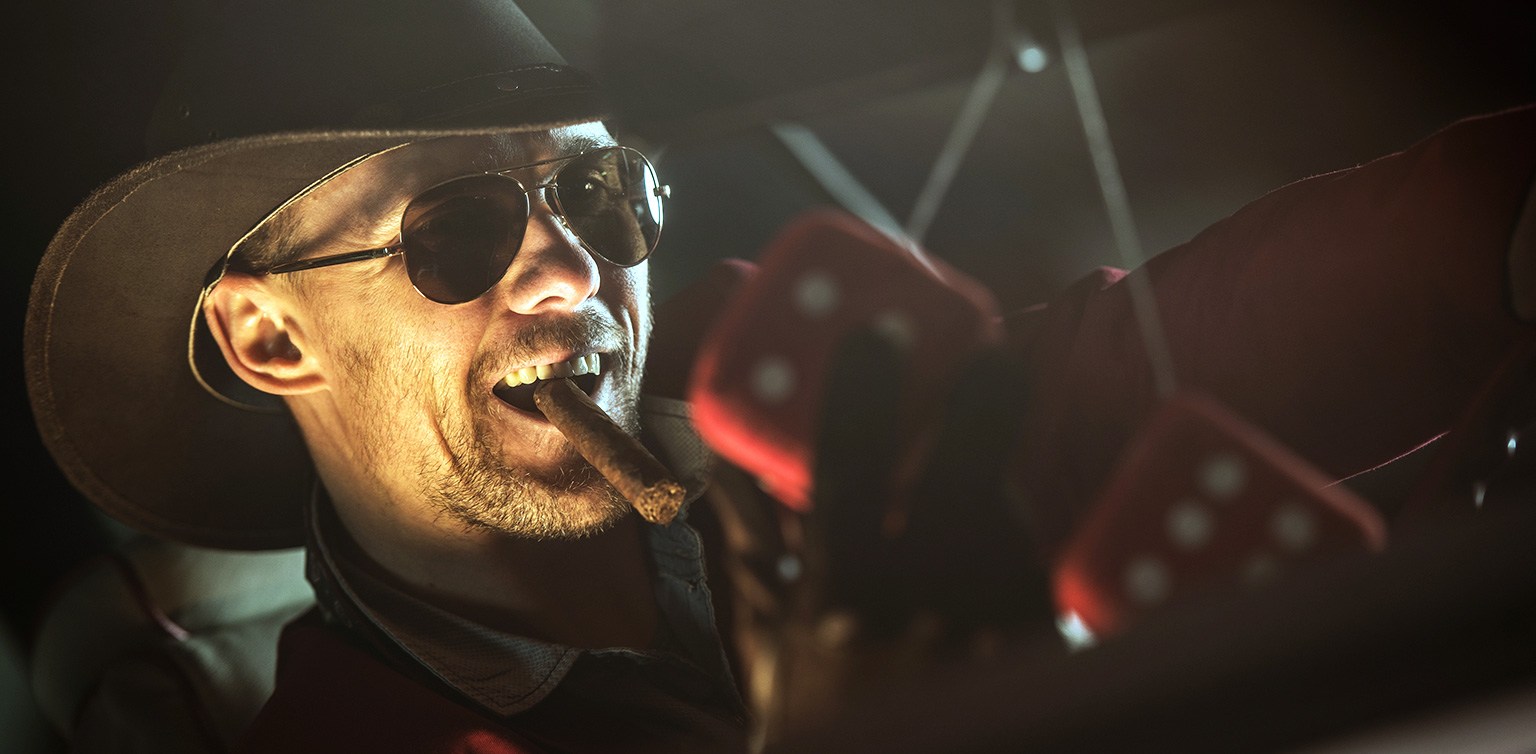 A photo of a man in a cowboy hat and sunglasses, smoking a cigar, with a pair of blurry red die hanging on his rear view mirror