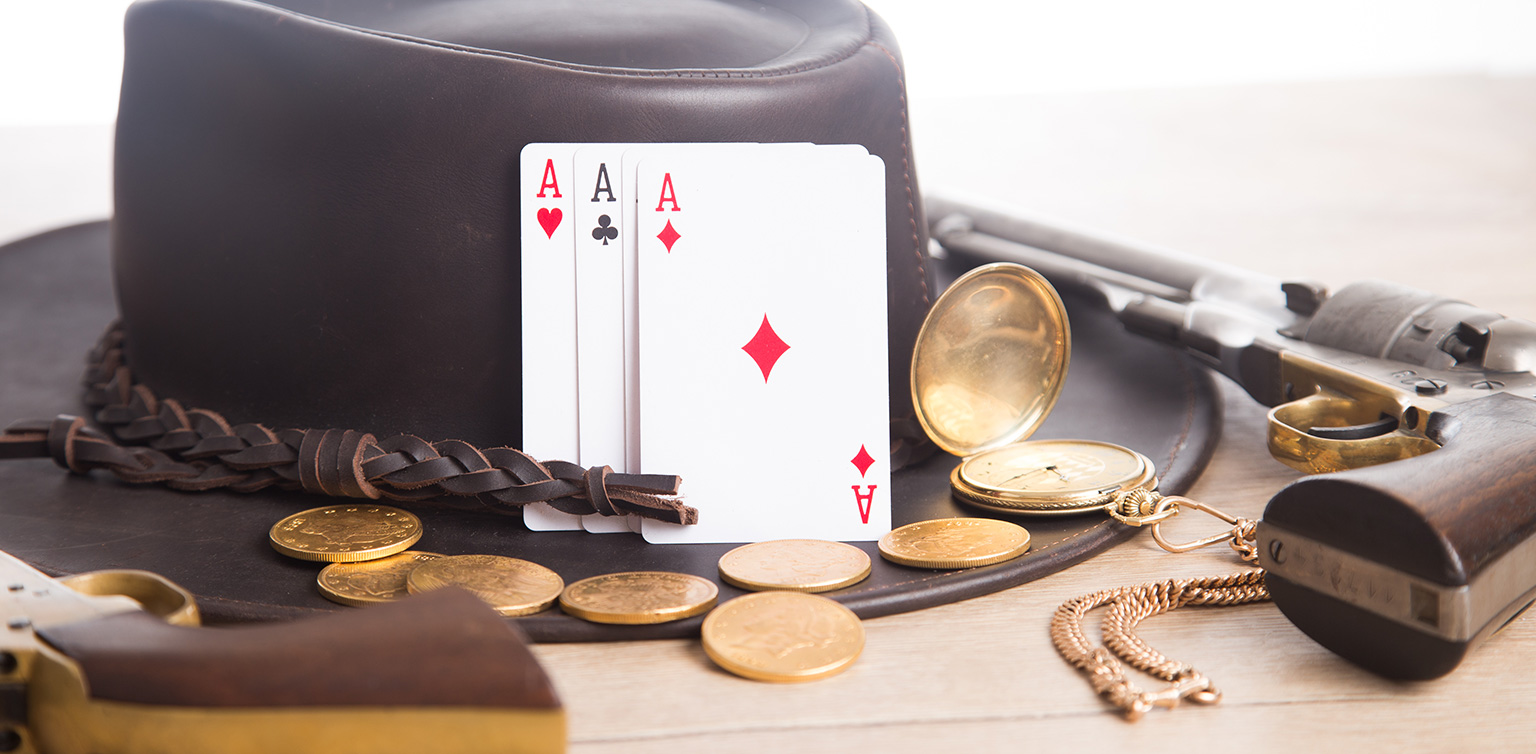 A photo of a cowboy hat with 3 aces leaning against it, surrounded by a pocket watch, handgun, gold coins on a wooden table